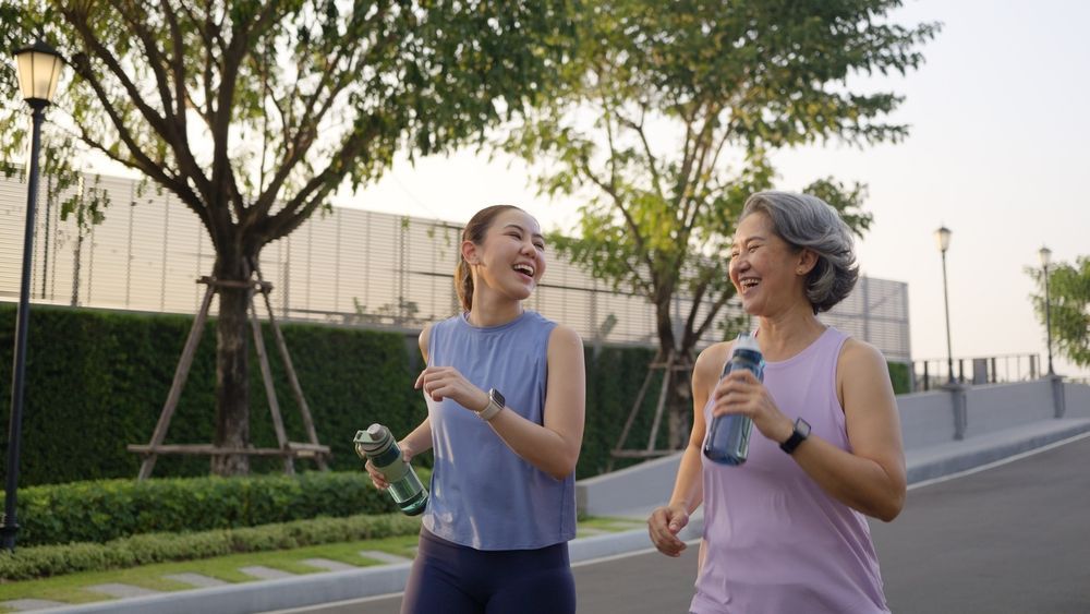 Two women jogging on a path, laughing. One holds a water bottle. Green foliage and lamp posts in the background.