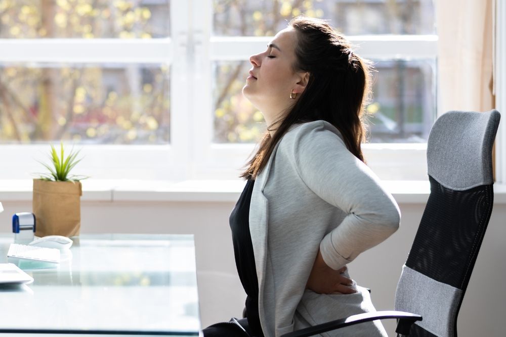Person sitting at a desk, leaning back and holding their lower back, suggesting back pain.