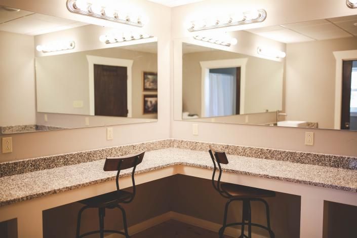 Bathroom vanity with large mirror, granite countertop, and two black stools under bright lights