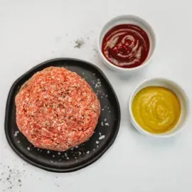 Raw ground meat with two small bowls of red and yellow sauces on a white surface