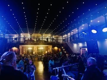 Dimly lit indoor concert venue with blue string lights and a crowd gathered on the floor and balcony.