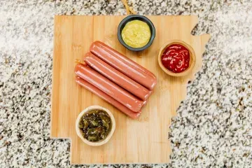 Hot dogs with three small bowls of condiments on a wooden cutting board on a granite counter