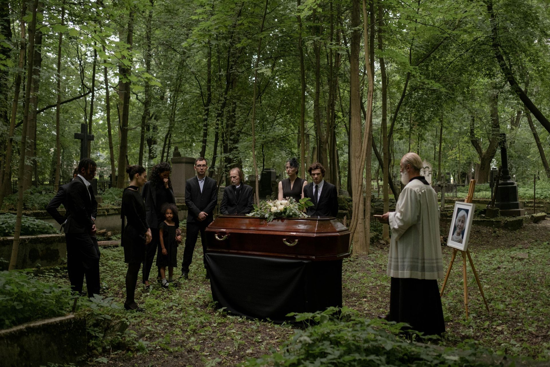 A group of people in black attire stand around a wooden coffin in a wooded cemetery while a priest leads a funeral.