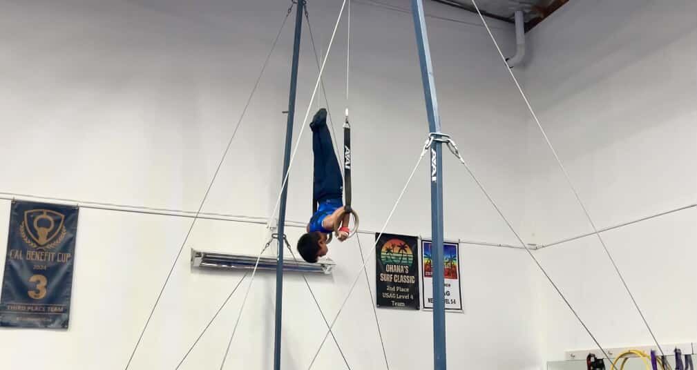 Gymnast in mid-air on gymnastic rings, body inverted, in a gym with banners and equipment.