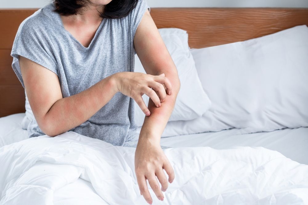 A woman is sitting on a bed scratching her arm.