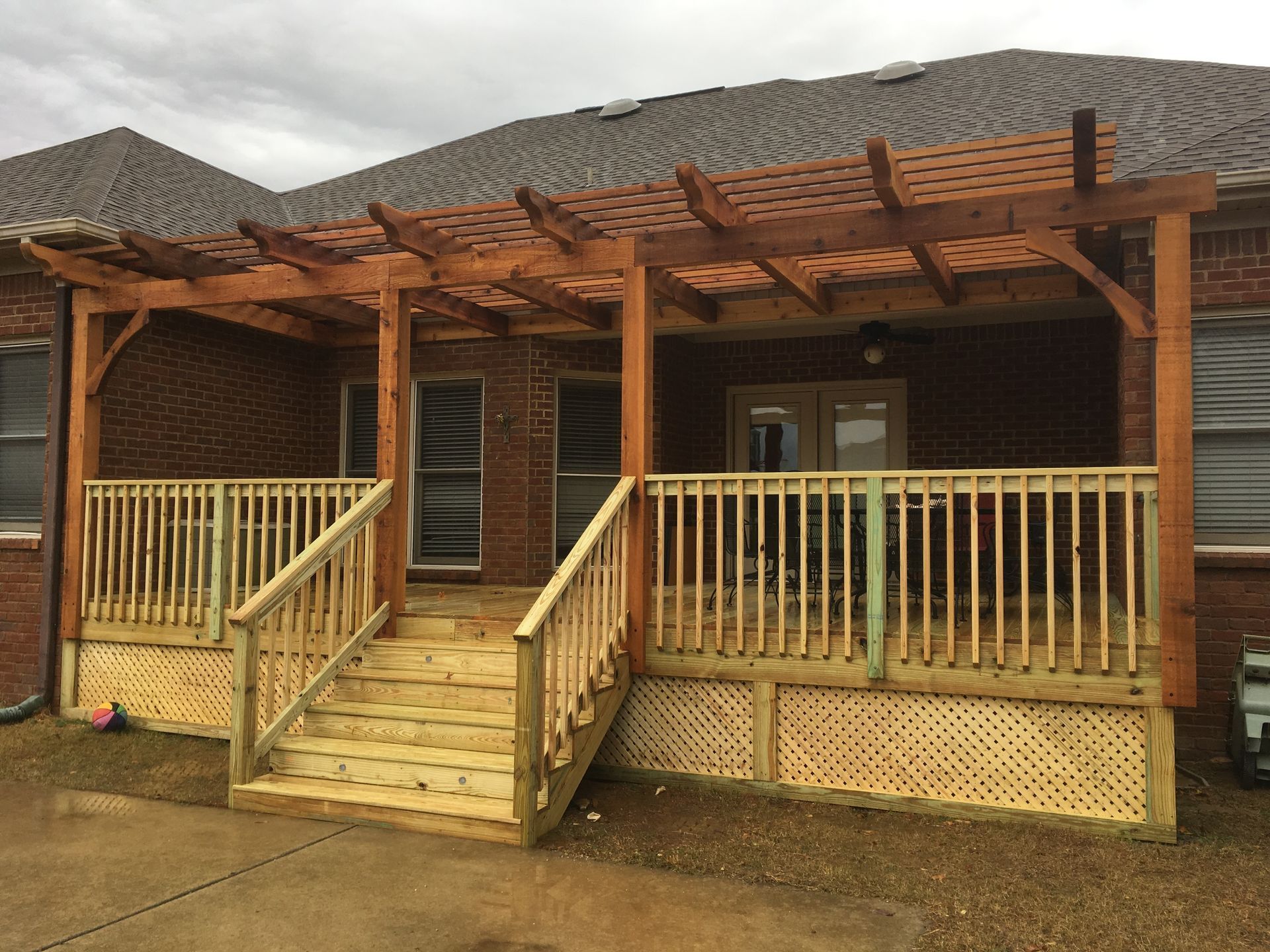 Wooden deck attached to a beige house, with stairs leading down to a grassy yard. A pergola is over the deck.