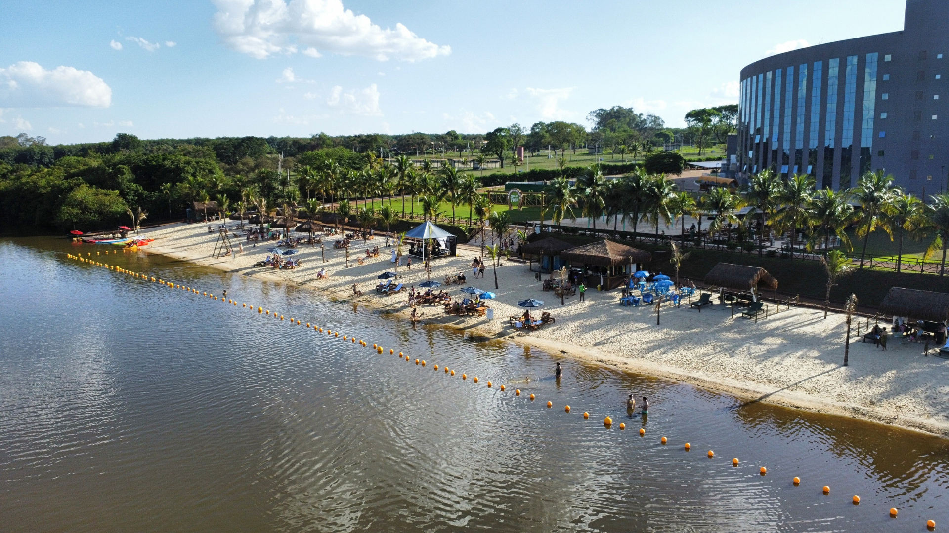 Praia de areia branca com pessoas relaxando. Árvores, lago e prédio moderno ao fundo.