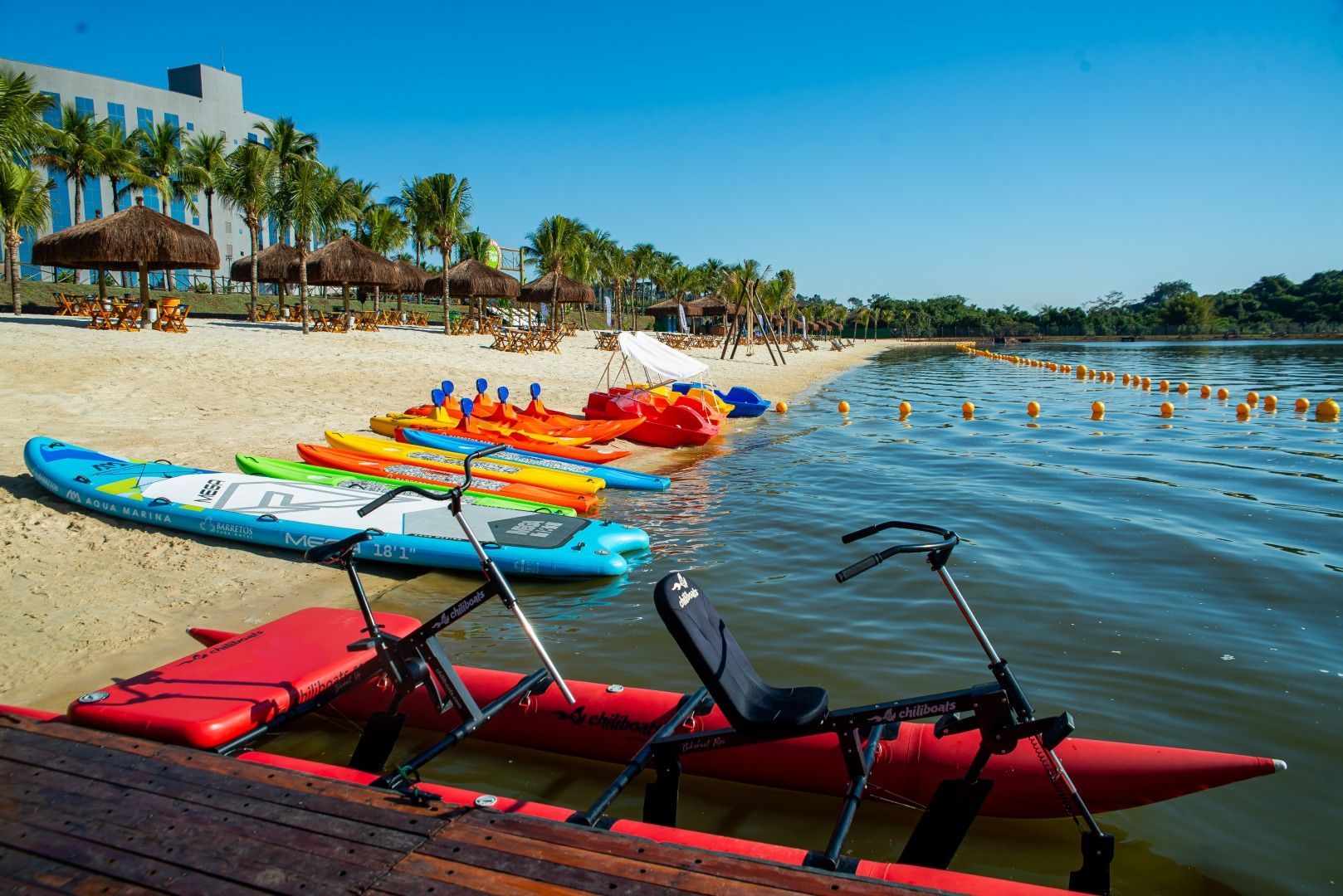 Embarcações enfileiradas em uma praia de areia. Céu azul brilhante. Pranchas de stand-up paddle, caiaques e pedalinhos. Palmeiras e um prédio ao fundo.