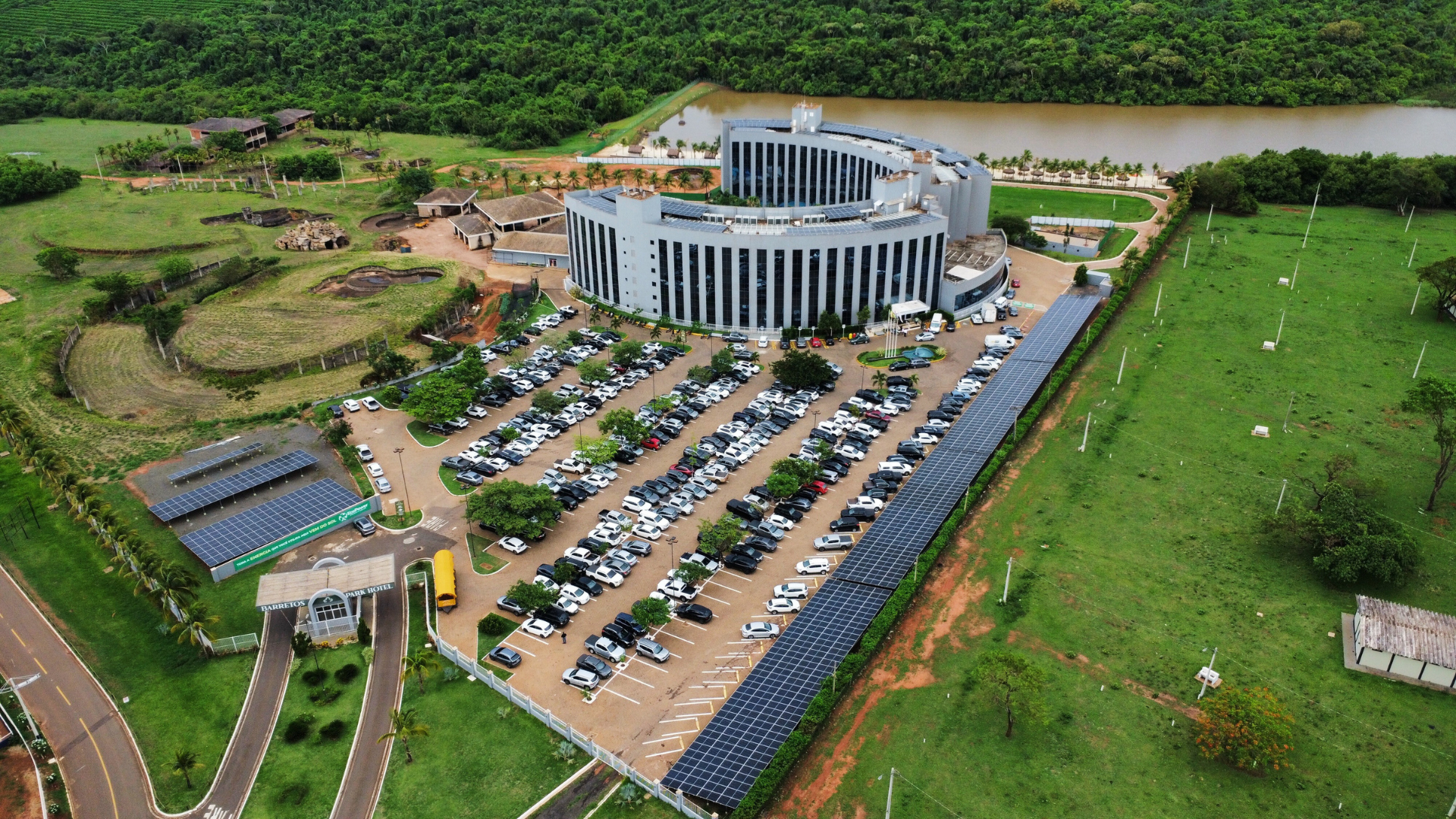 Vista aérea de um grande edifício cinza e curvo com amplo estacionamento, campos verdes e um rio ao fundo.