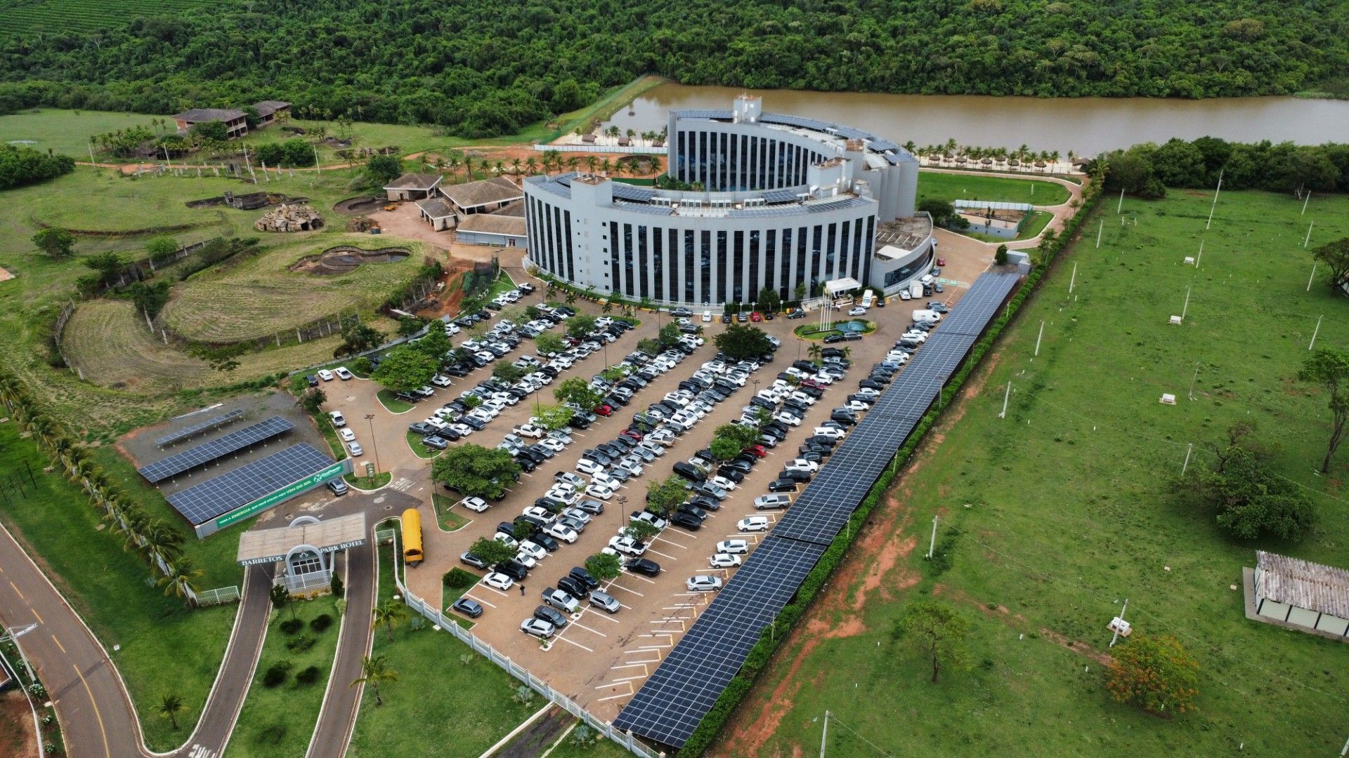 Vista aérea de um grande edifício cinza curvo com estacionamento e painéis solares, inserido em uma paisagem verde.
