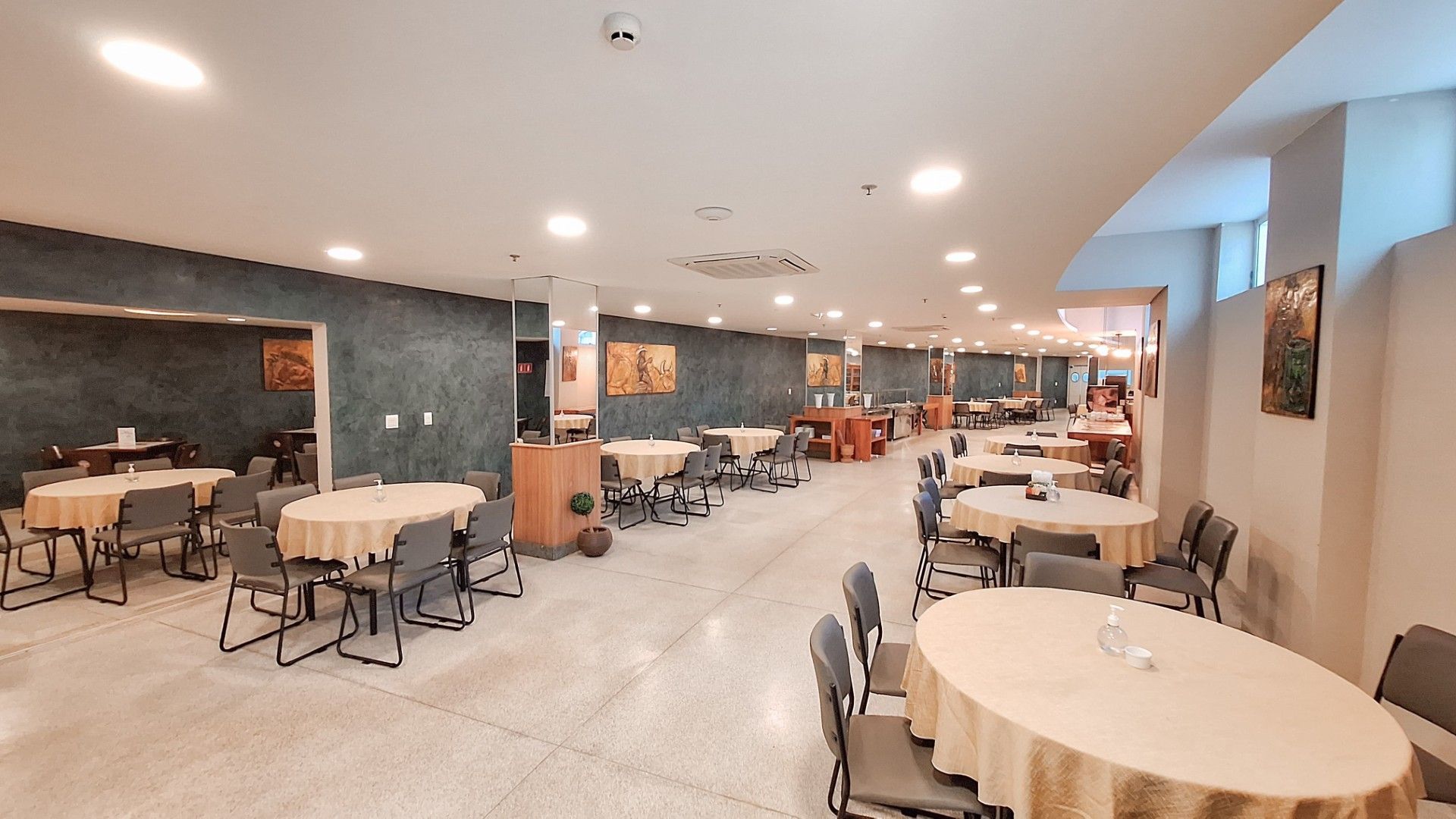 Interior espaçoso de restaurante com mesas redondas cobertas com toalhas de mesa bege e cadeiras cinza.