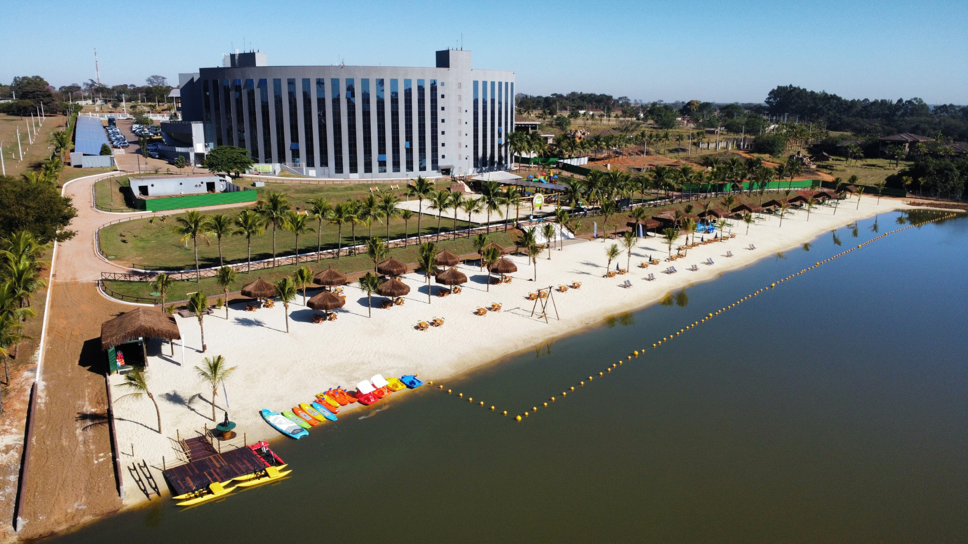 Praia à beira do lago com guarda-sóis e caiaques. Grande edifício moderno ao fundo. Dia ensolarado.