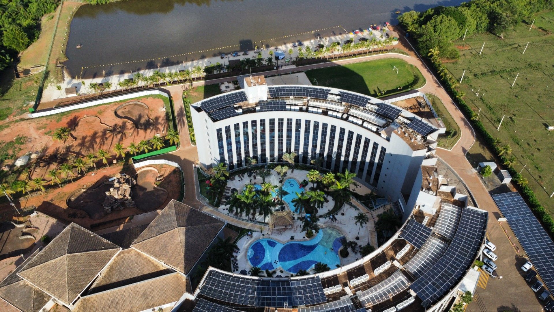 Vista aérea de um edifício de hotel curvo com piscina e lago, rodeado por árvores e campos.