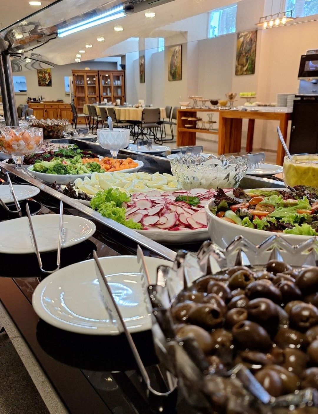 Mesa de buffet com diversas saladas e azeitonas. Ambiente interno com mesas e cadeiras.