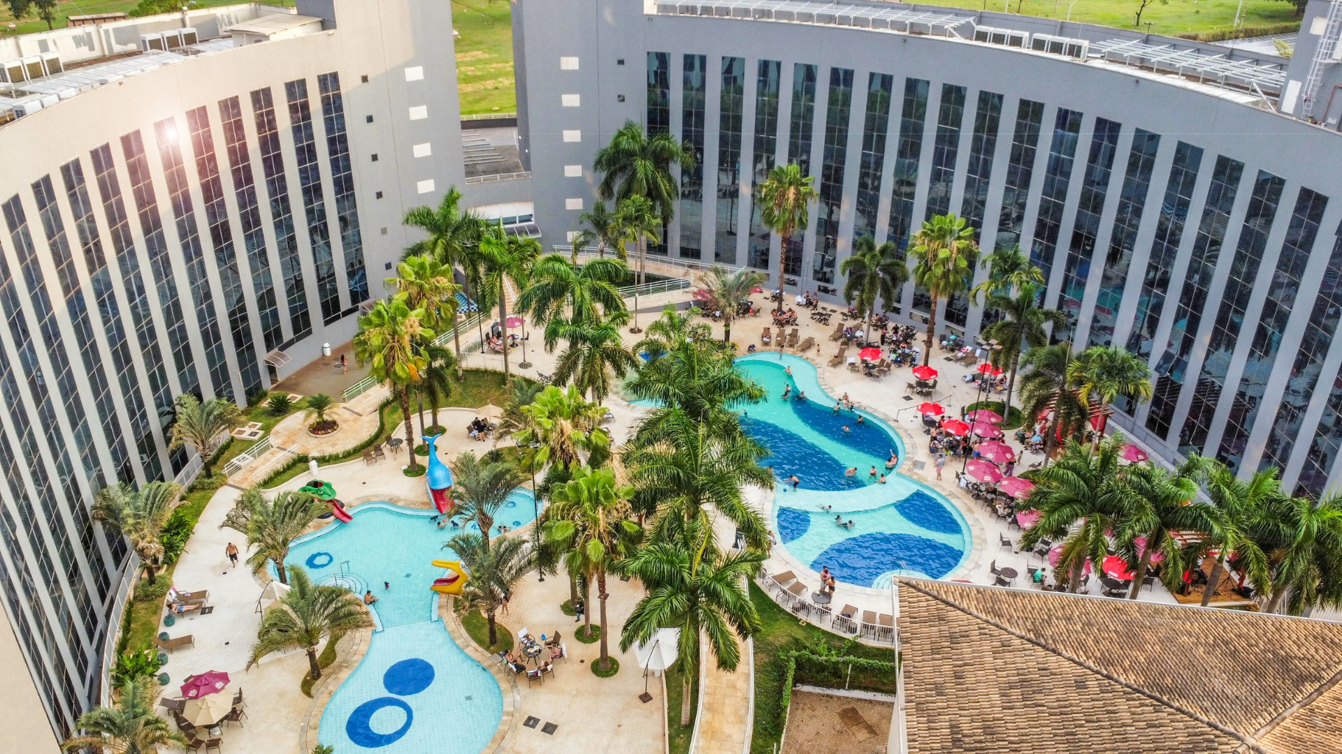 Vista aérea de um resort com edifícios curvos, várias piscinas, palmeiras e pessoas.