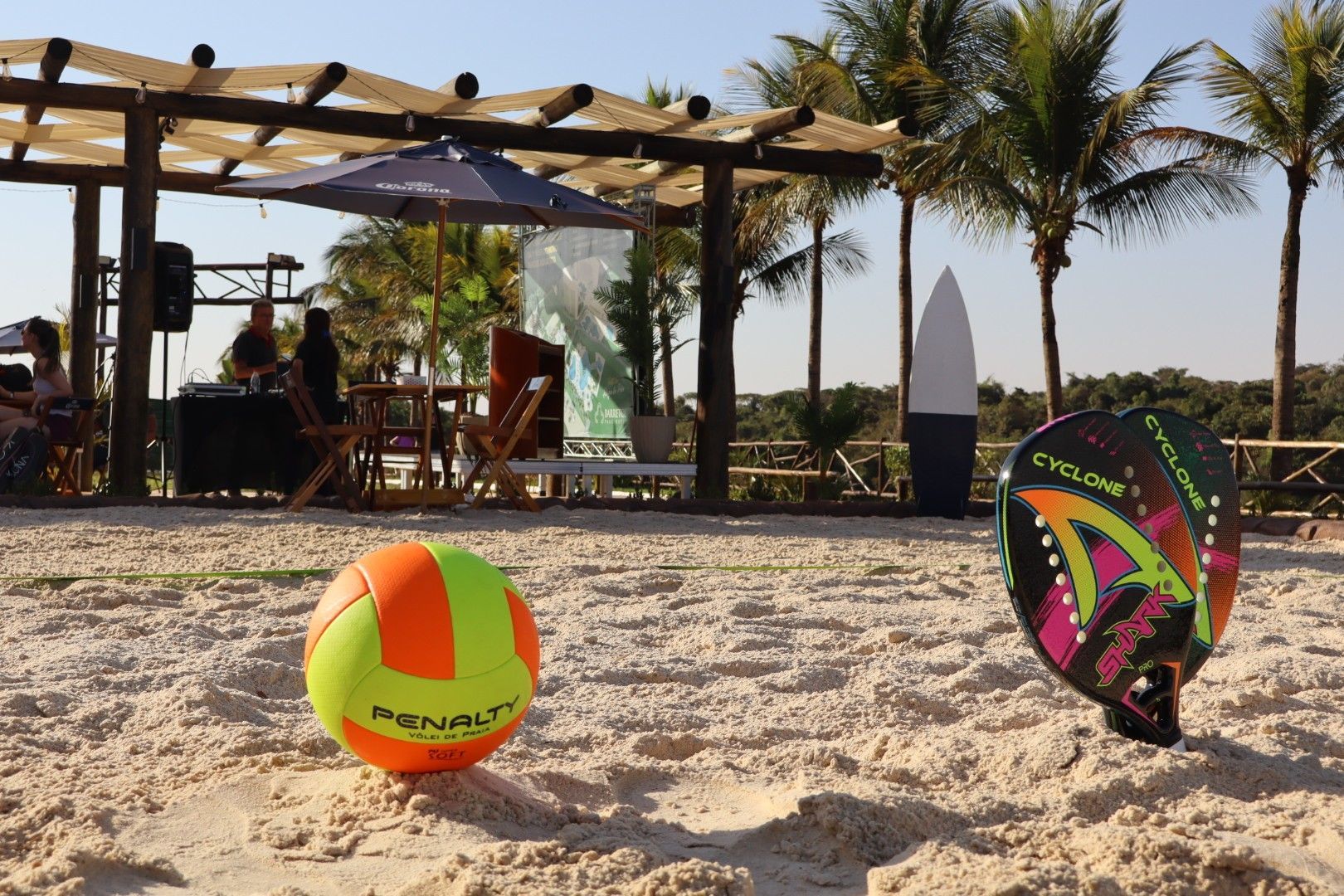 Vôlei de praia e frescobol na areia. Uma tenda, pessoas e palmeiras ao fundo. Dia ensolarado.