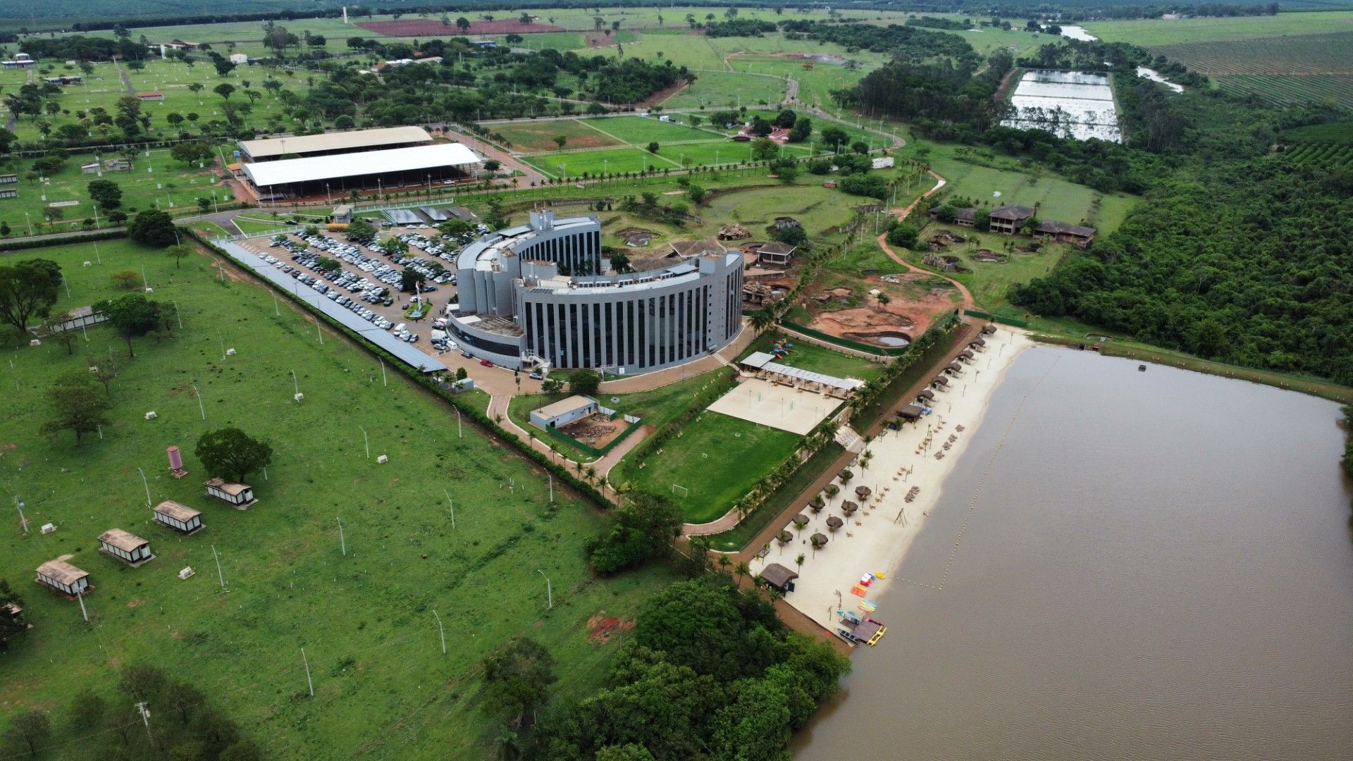 Vista aérea: um complexo de edifícios modernos com praia e lago, rodeado por campos verdes e árvores.