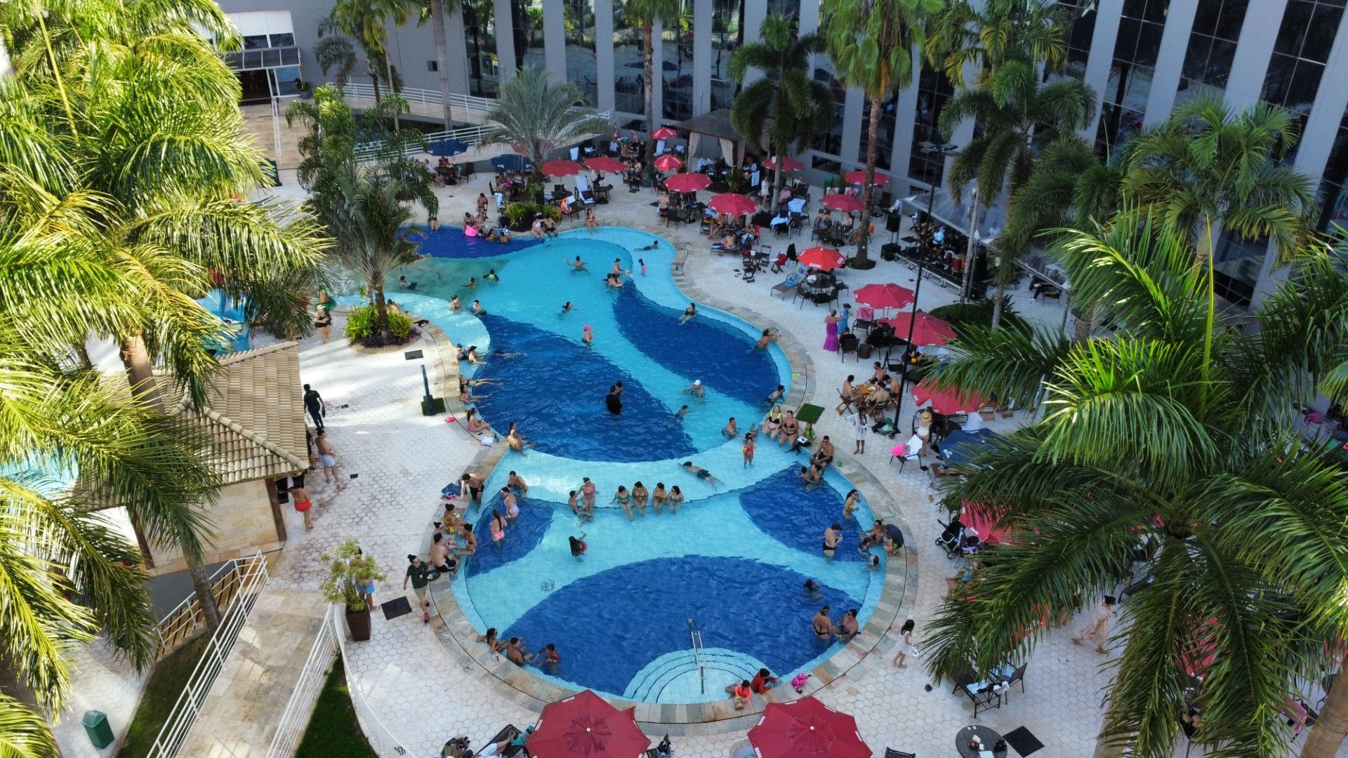 Vista aérea de uma piscina lotada com pessoas nadando, cercada por palmeiras e mesas ao ar livre.