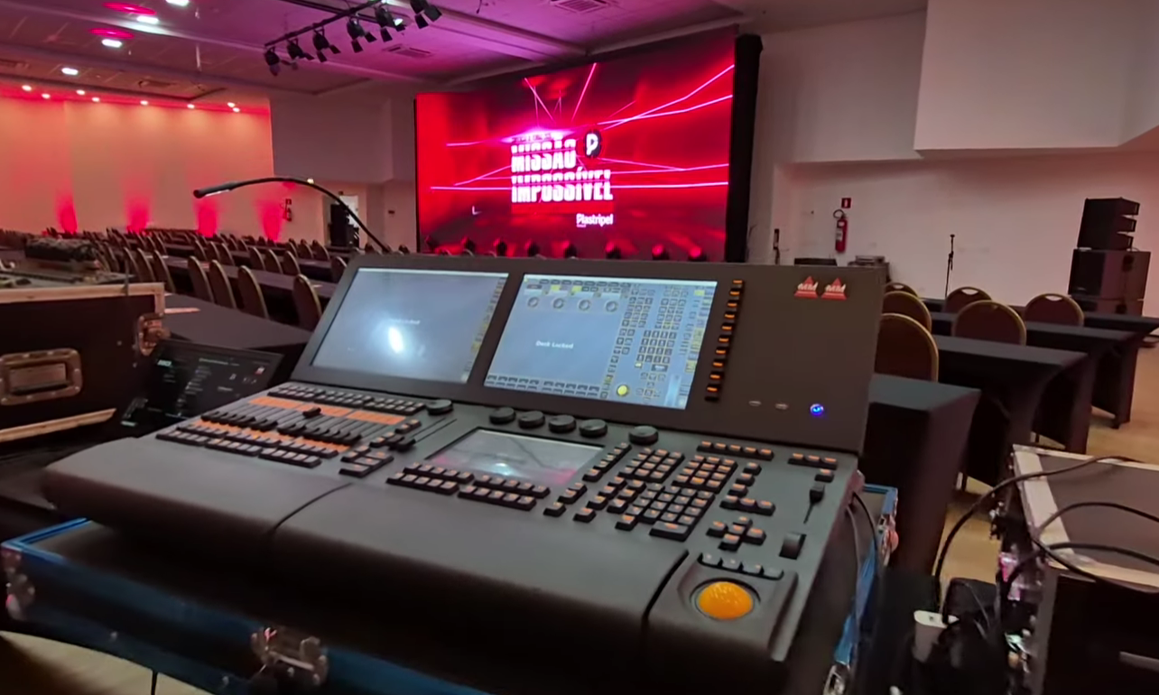 Elegante salão de eventos com mesas e cadeiras cobertas com toalhas vermelhas, árvore de Natal e palco sob um teto branco com luzes vermelhas e brancas.