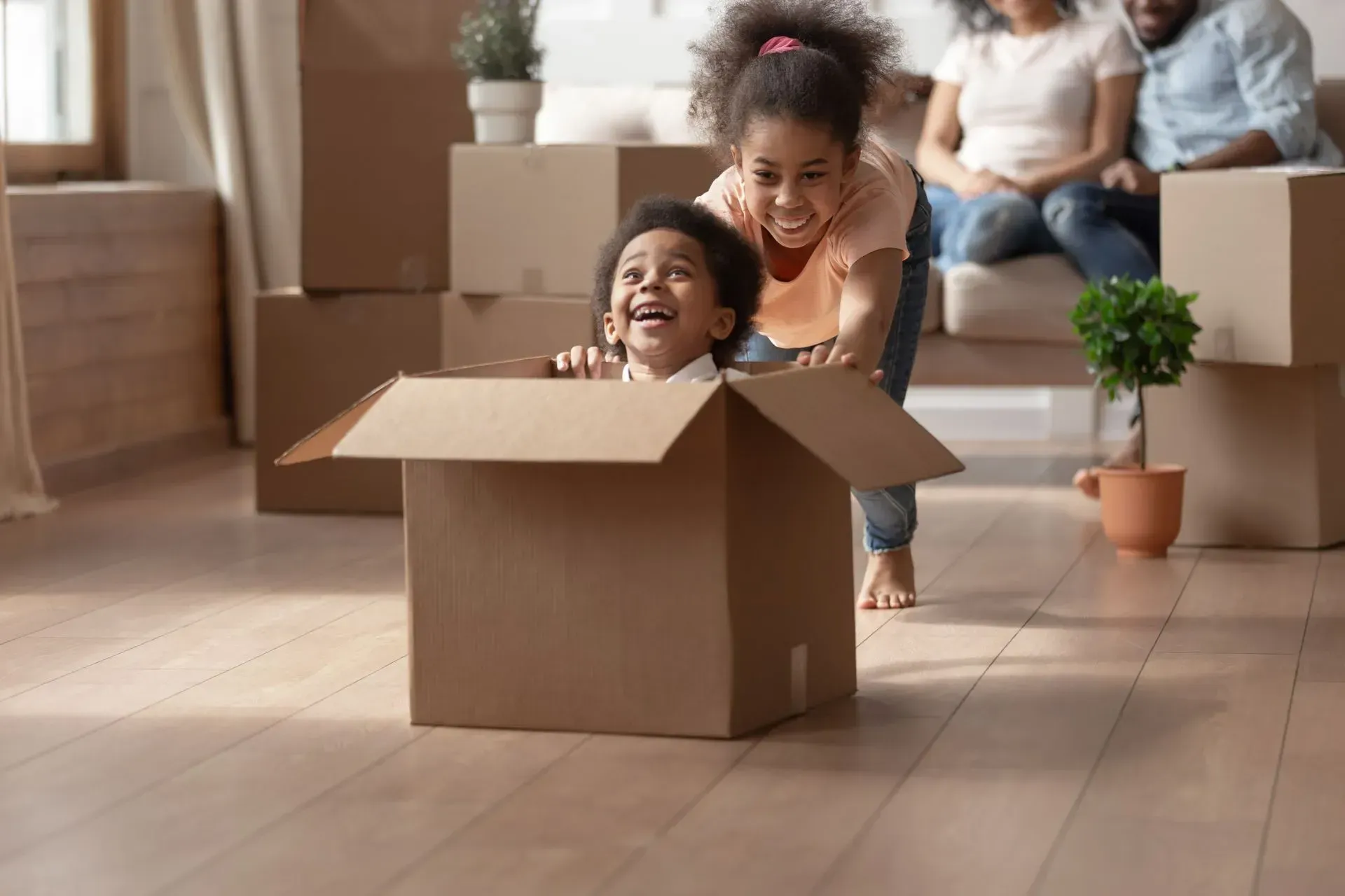 An older child pushes a younger child sitting inside a cardboard box in a living room filled with moving boxes.