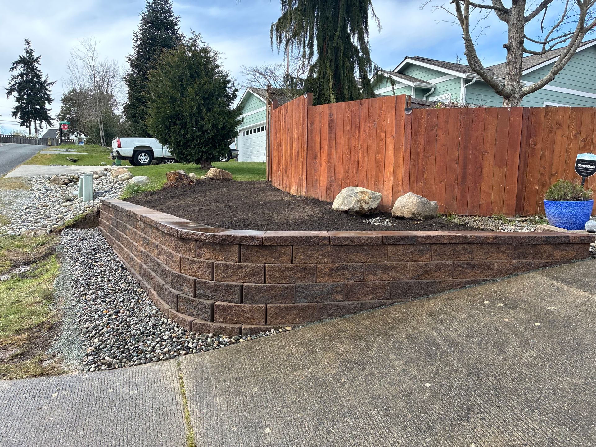 Retaining wall with brown fence and dirt planter beside a driveway in a suburban yard