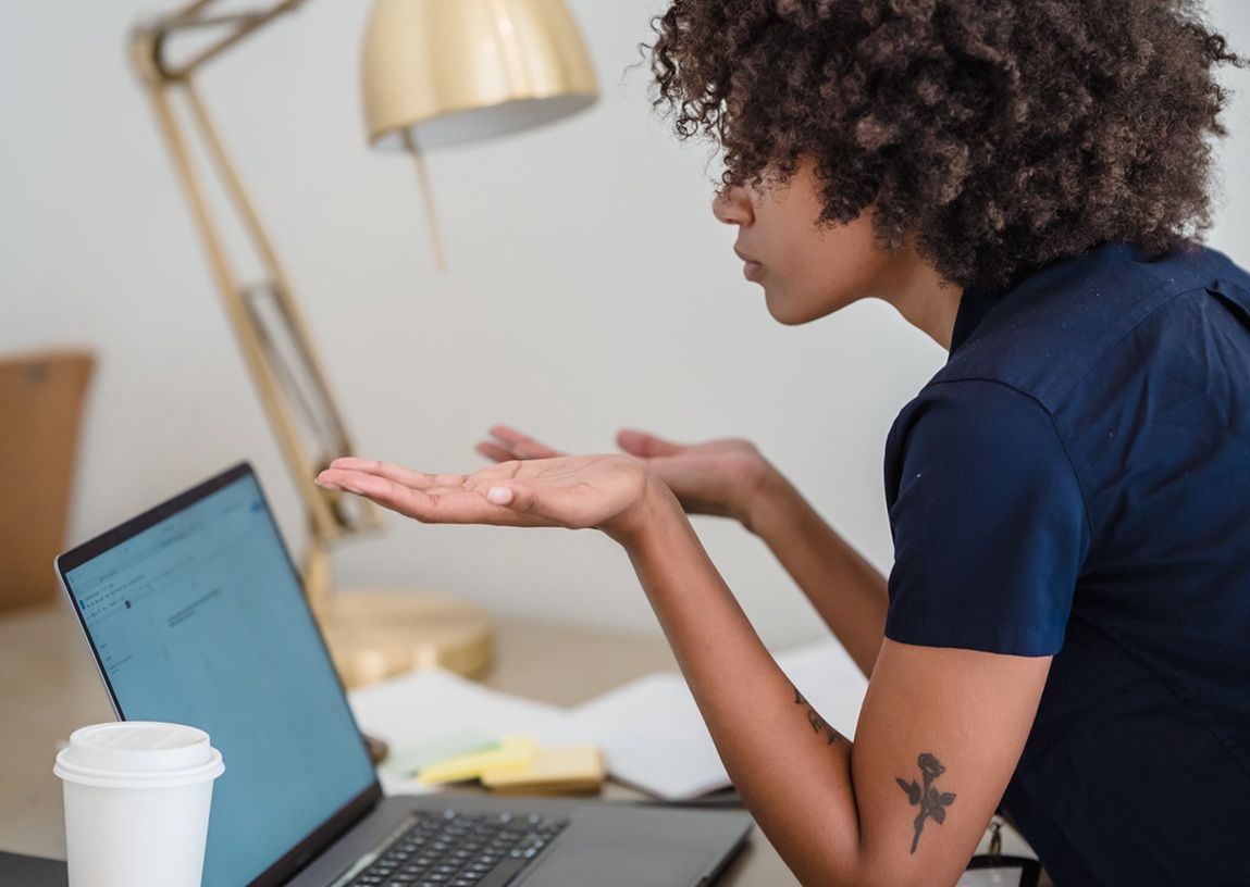 Person seated at a desk, gesturing while looking at a laptop in a home office