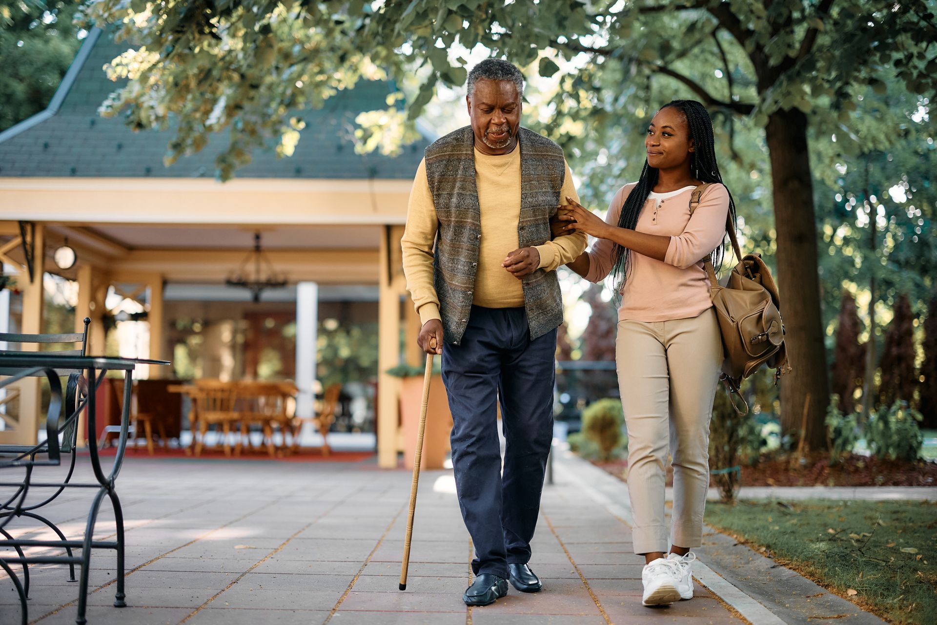 Woman helps man with a cane walk on a brick path outside a building.