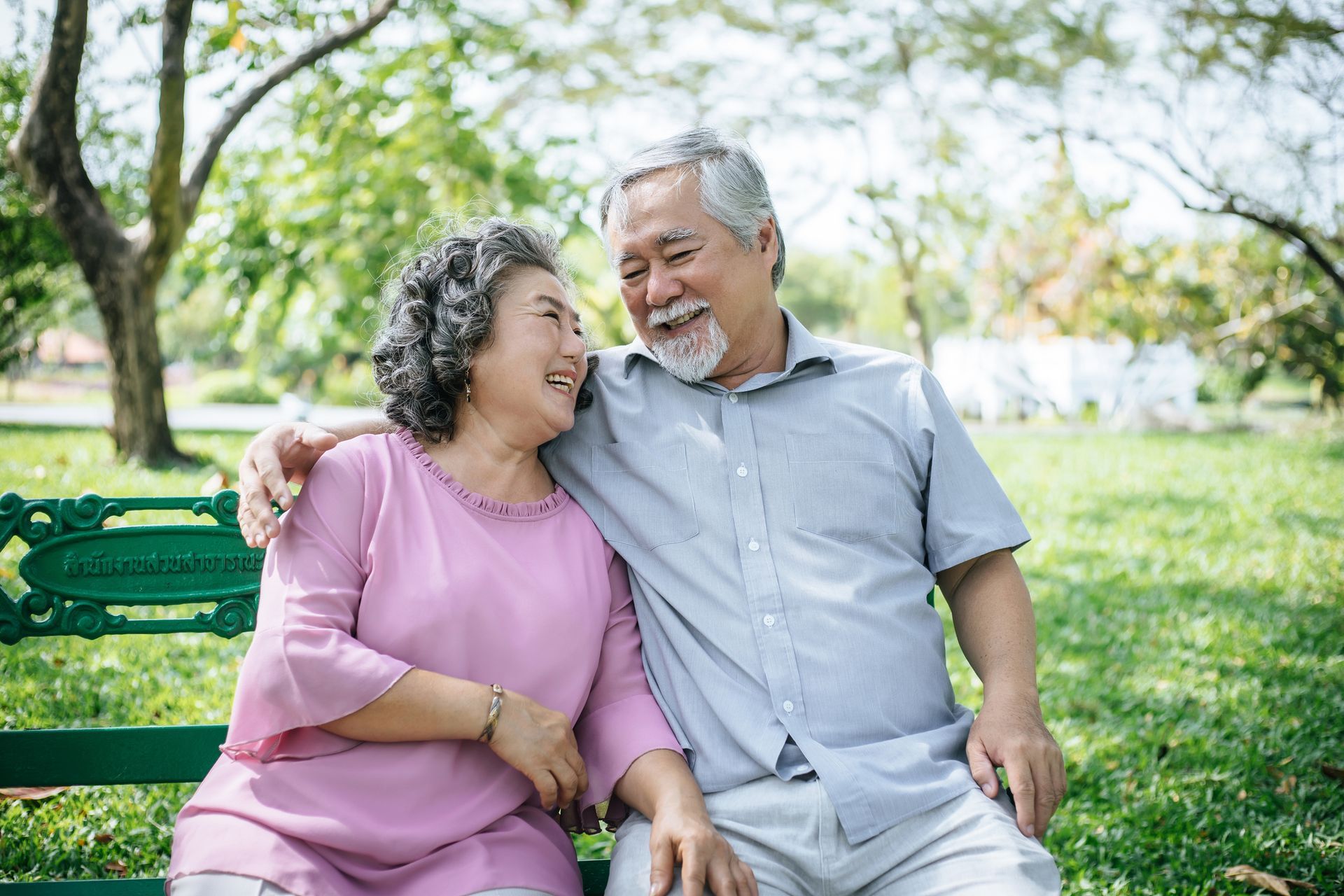 Elderly couple sitting on a park bench, smiling, embracing outdoors.