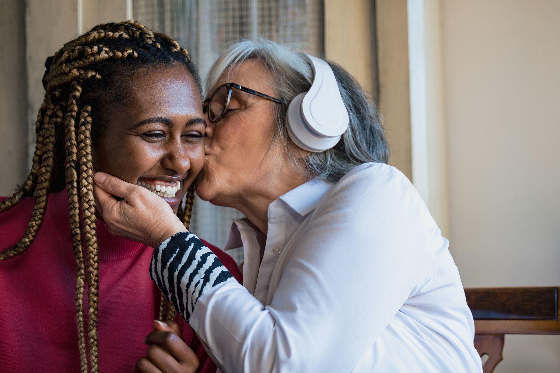 Woman kisses another woman on the cheek; both are smiling. One wears headphones.
