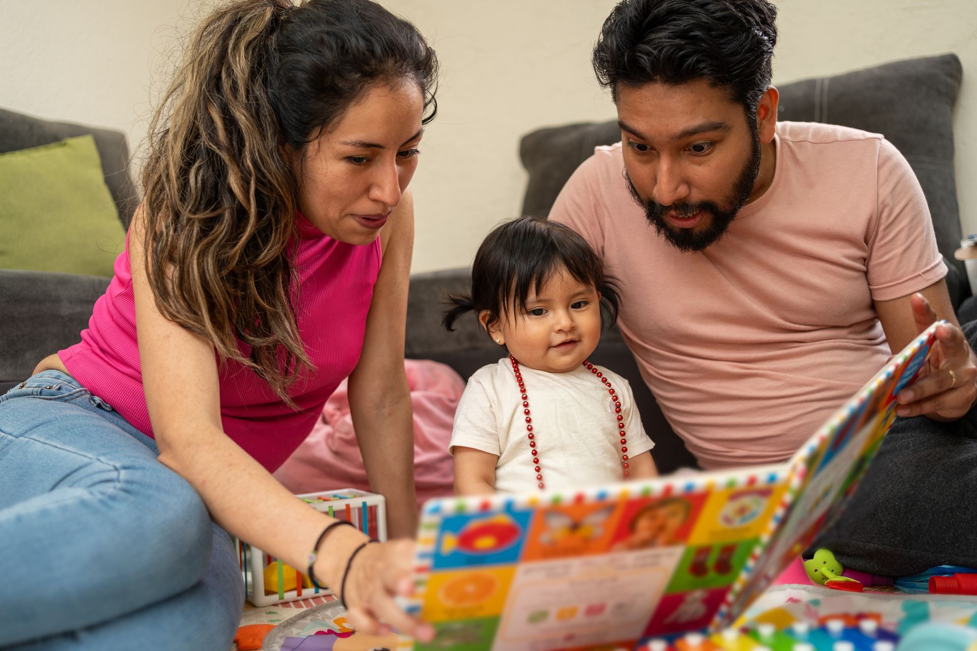 Family reading a book together on the floor. Parents lean over a child, looking at colorful pages.
