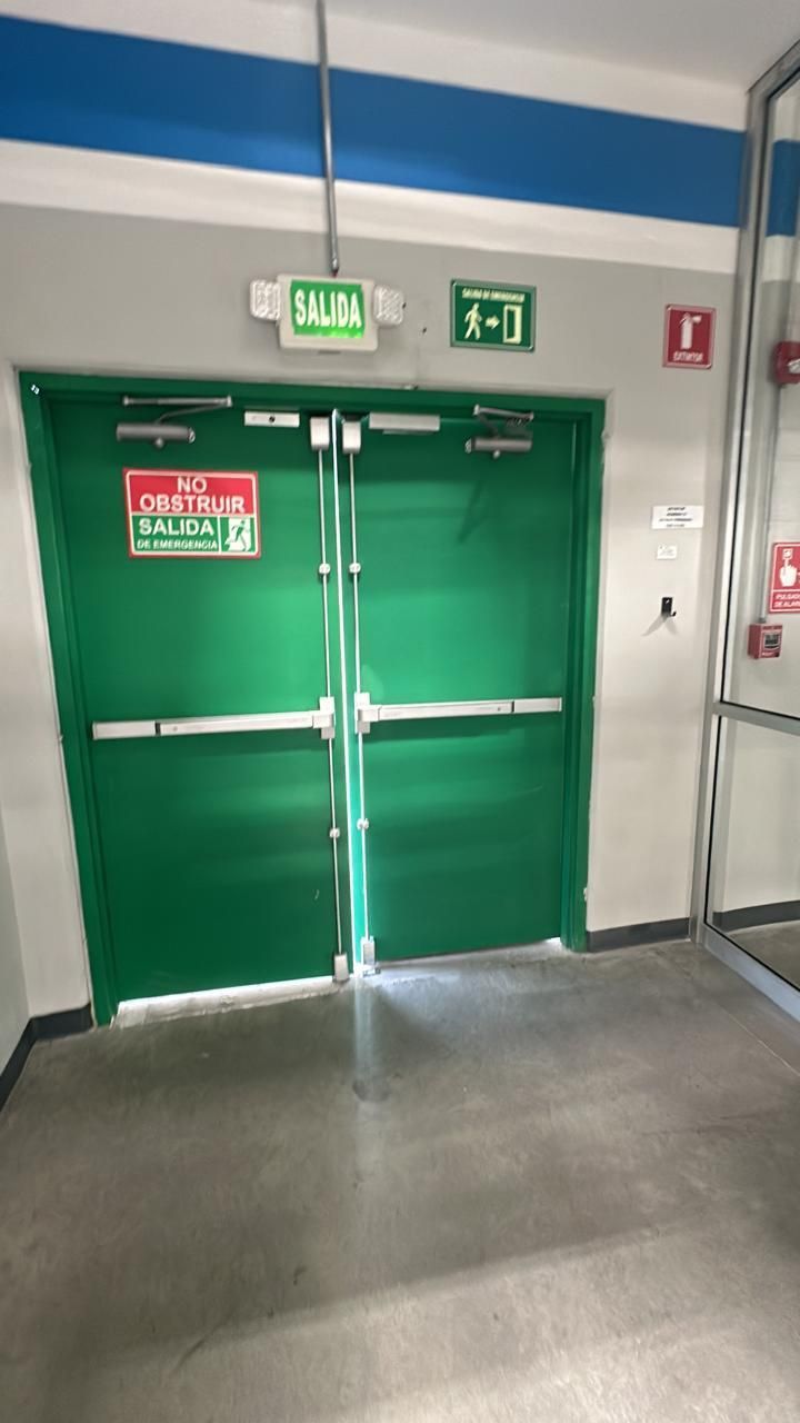 Puertas dobles verdes con señales de salida, barras antipánico y botón de alarma contra incendios.