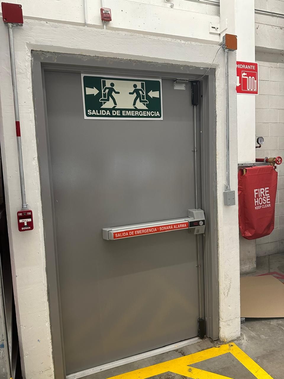 Puerta de salida de emergencia con barra antipánico, señal de salida verde, alarma contra incendios y manguera contra incendios.