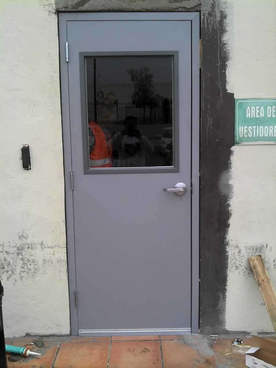 Puerta metálica gris con ventana, situada en una pared de color claro, con un cartel en español cerca.