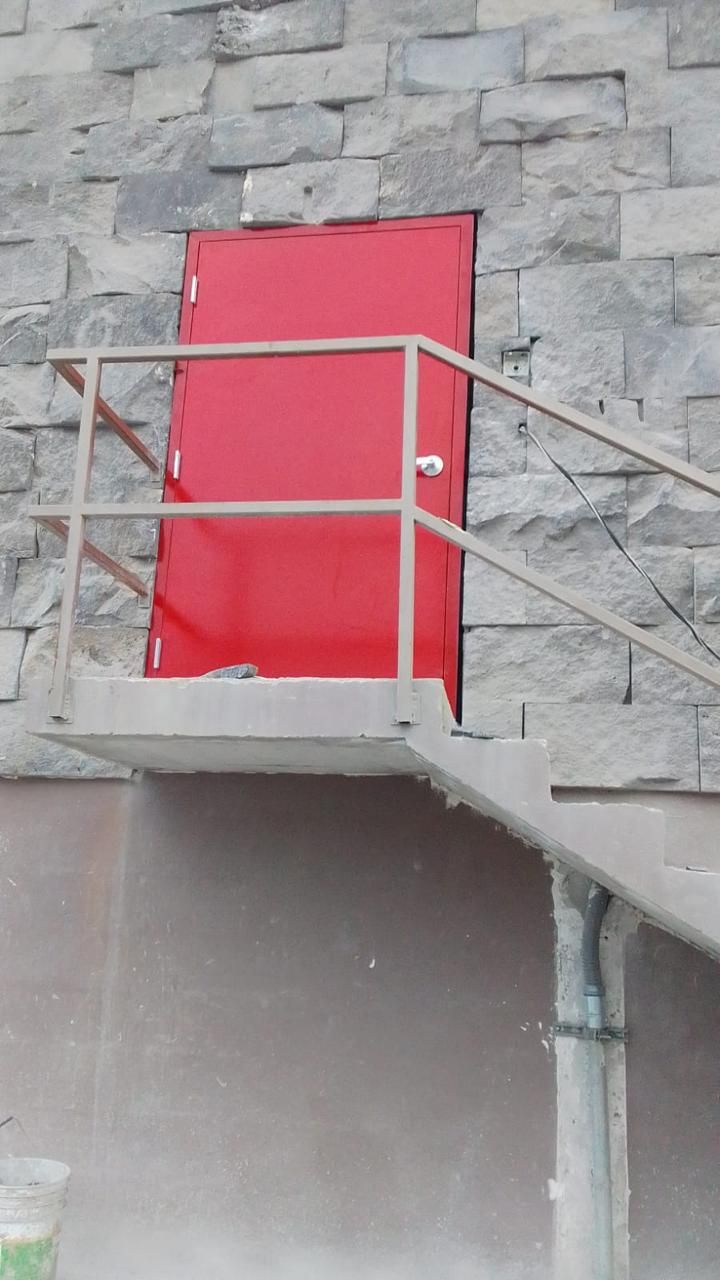 Puerta roja empotrada en la pared de piedra, con escaleras y barandilla metálicas que conducen a ella.