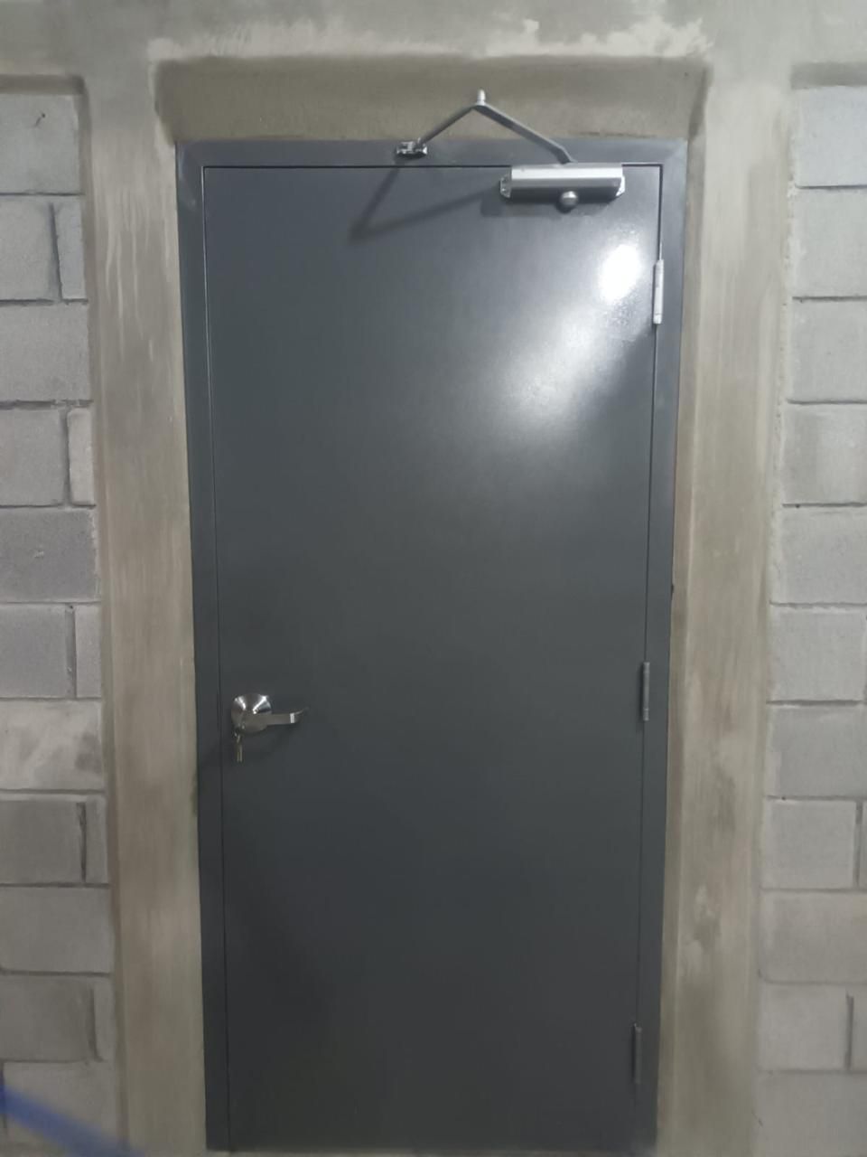 Puerta metálica gris con cierrapuertas en una pared de bloques de hormigón.