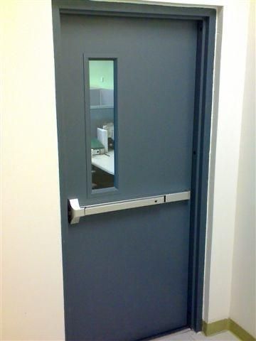 Puerta gris con ventana rectangular y barra de salida de emergencia en una habitación de paredes blancas.