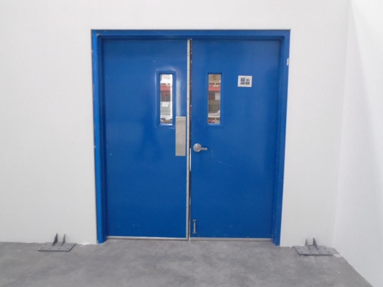 Puertas dobles azules con pequeñas ventanas y manilla y herrajes plateados, enmarcadas en una pared blanca.