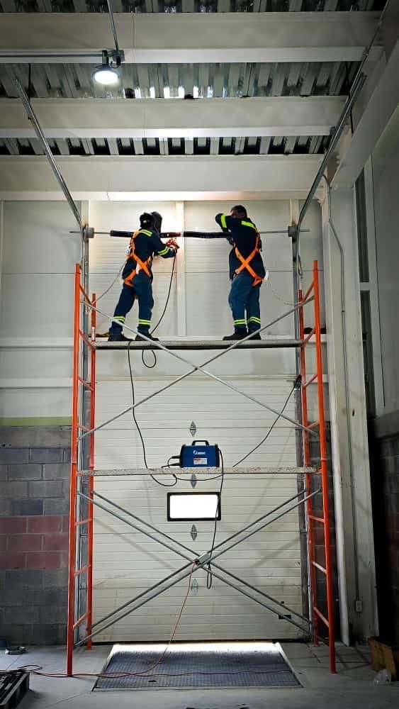 Dos trabajadores con equipo de seguridad en un andamio, inspeccionando una puerta de garaje cerrada en un entorno industrial.