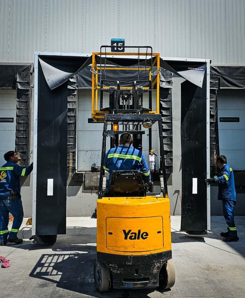 Operador de montacargas en un muelle de carga, con trabajadores ajustando el sello del muelle. Montacargas amarillo, uniformes azules.