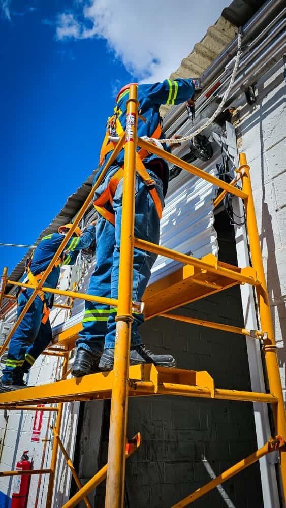 Dos trabajadores con equipo de seguridad sobre un andamio amarillo reparando el exterior de un edificio, cielo azul brillante.