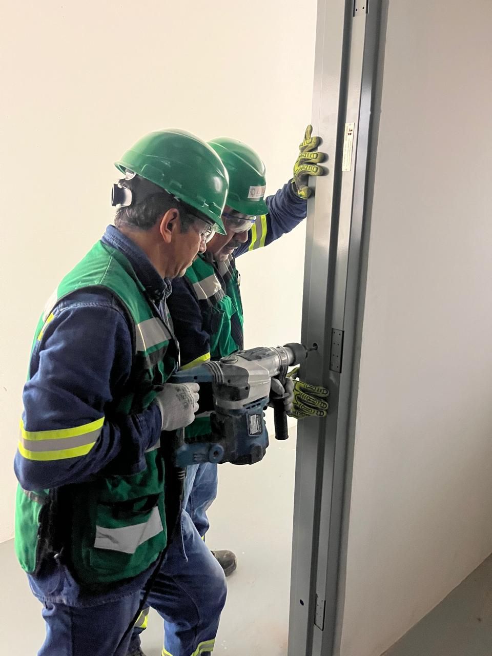 Dos trabajadores con cascos y chalecos antibalas taladran el marco metálico de una puerta en una habitación blanca.