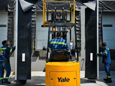 Operador de montacargas en un muelle de carga; dos trabajadores guían grandes paneles. Montacargas amarillo, uniformes azules.