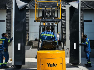 Operador de montacargas en un muelle de carga; dos trabajadores guían grandes paneles. Montacargas amarillo, uniformes azules.