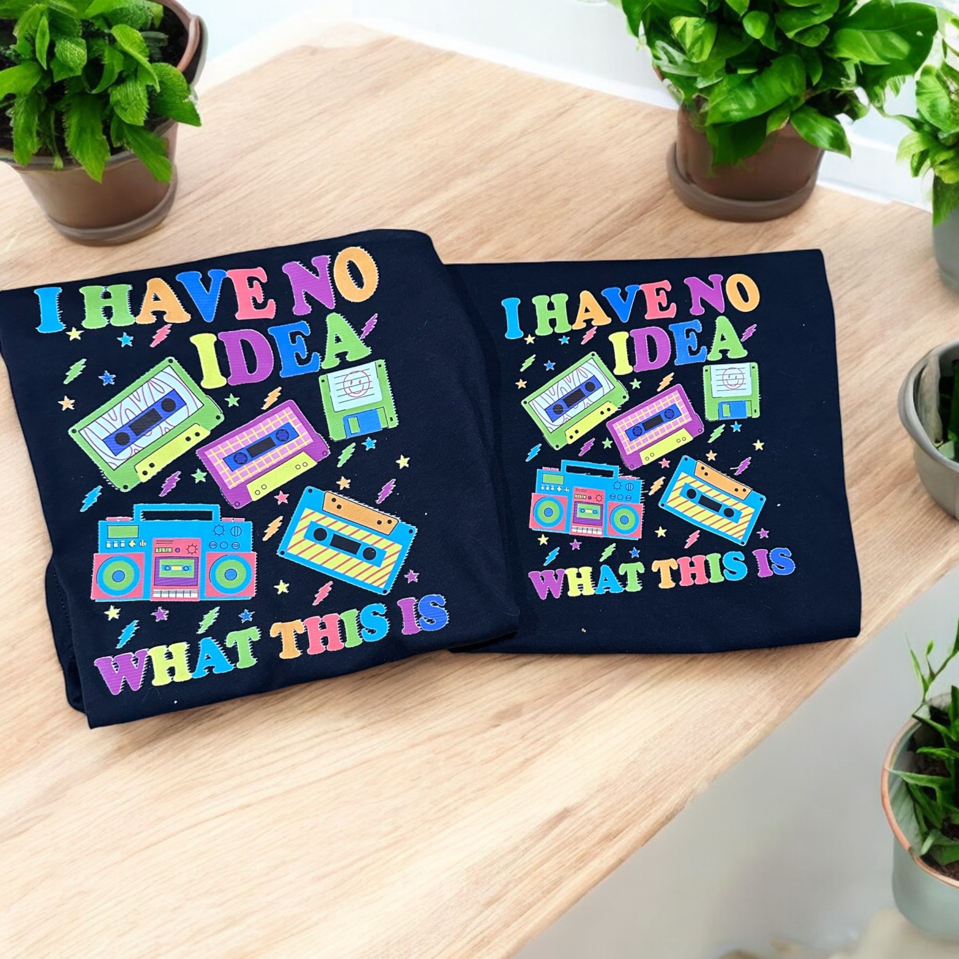 Two navy blue t-shirts with colorful "I HAVE NO IDEA WHAT THIS IS" retro design, surrounded by plants on a wooden surface.