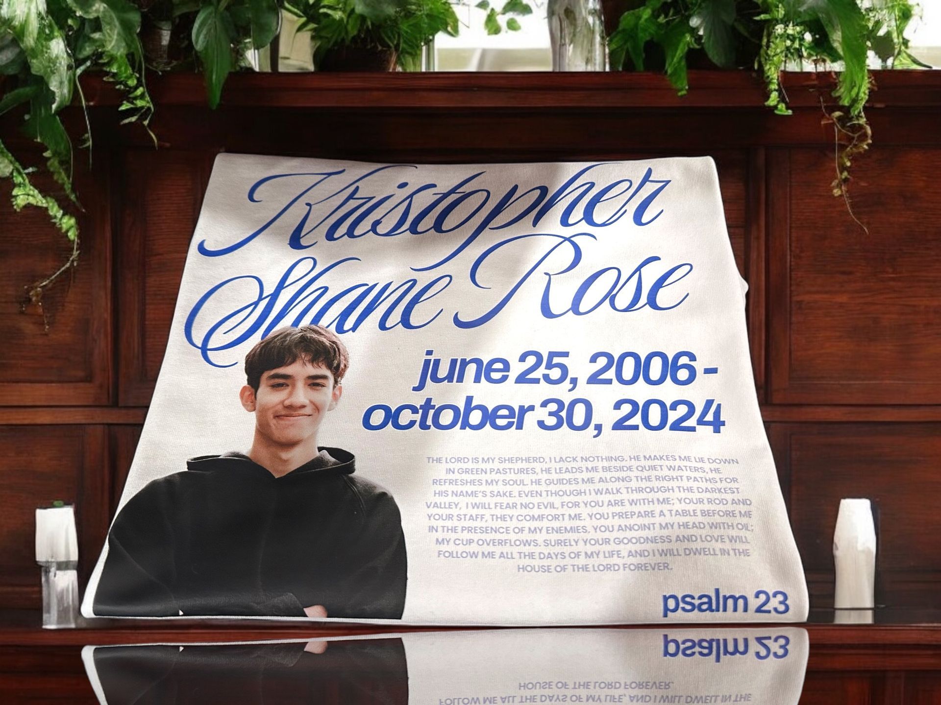Memorial poster for Kristopher Shane Rose, featuring a photo of him, dates, and Psalm 23, displayed on a dark wooden surface.