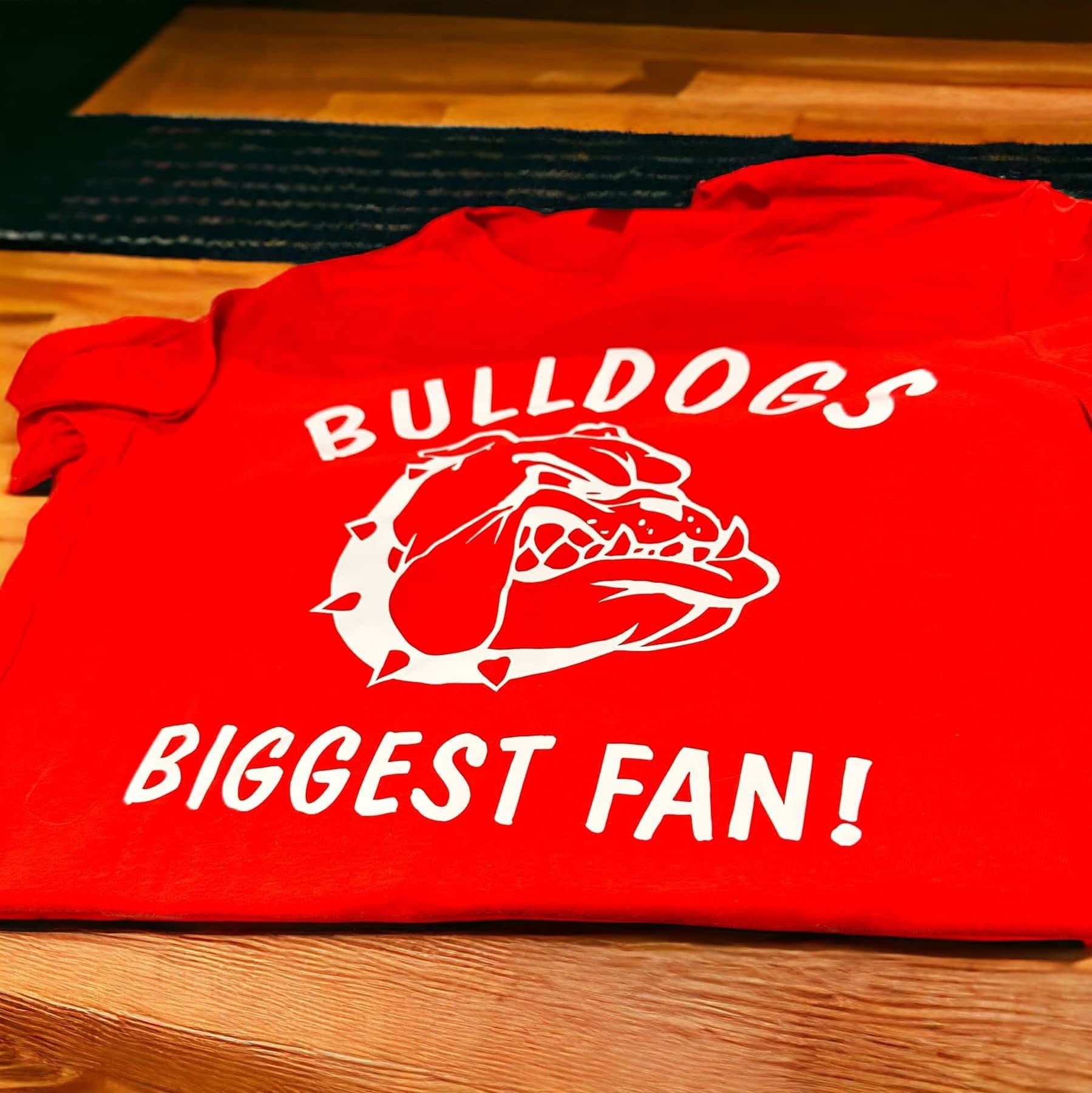 Red "Bulldogs Biggest Fan!" t-shirt with bulldog graphic on a wooden surface.