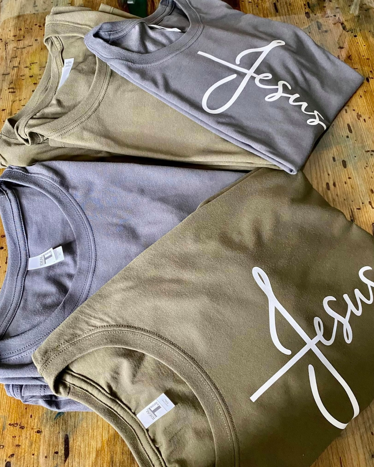 Pile of folded t-shirts in olive, gray, and tan, each with "Jesus" in white script.