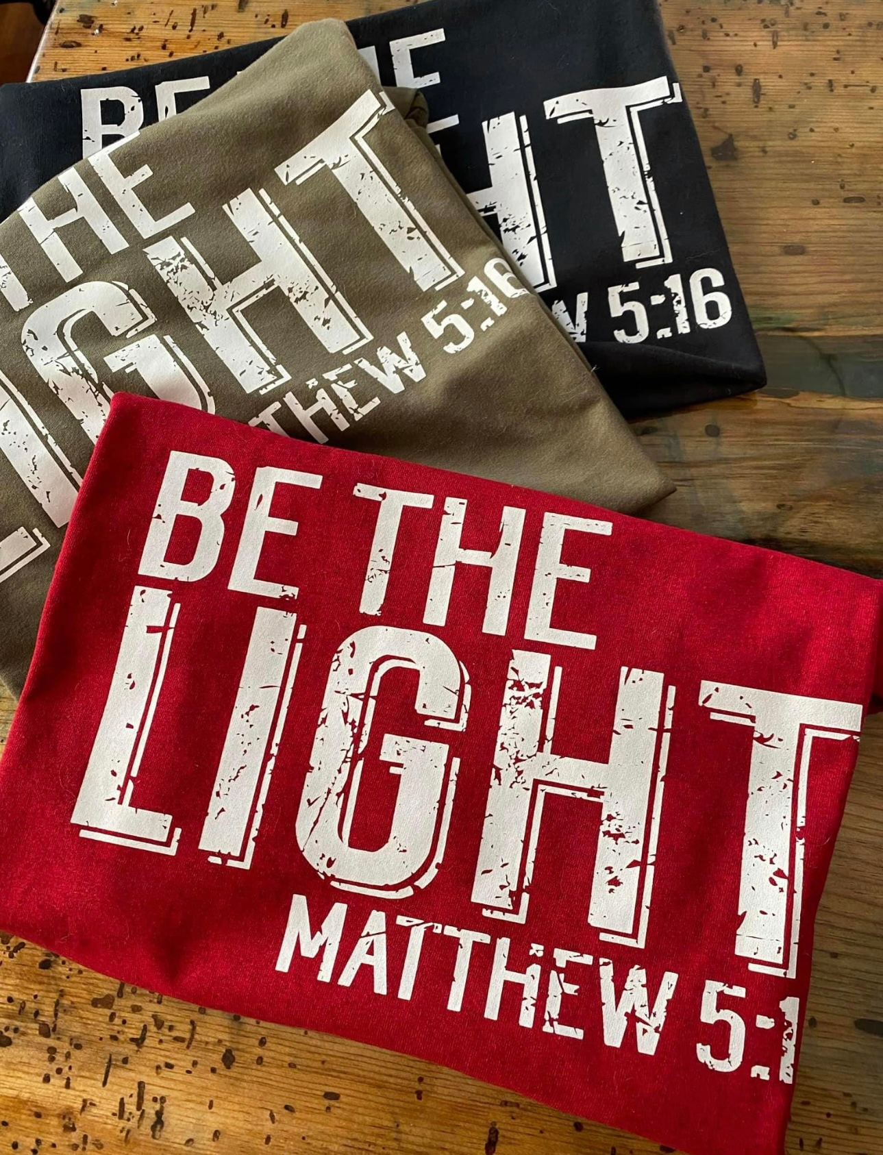 Folded t-shirts in red, brown, black with "BE THE LIGHT" and "Matthew 5:16" printed in white.