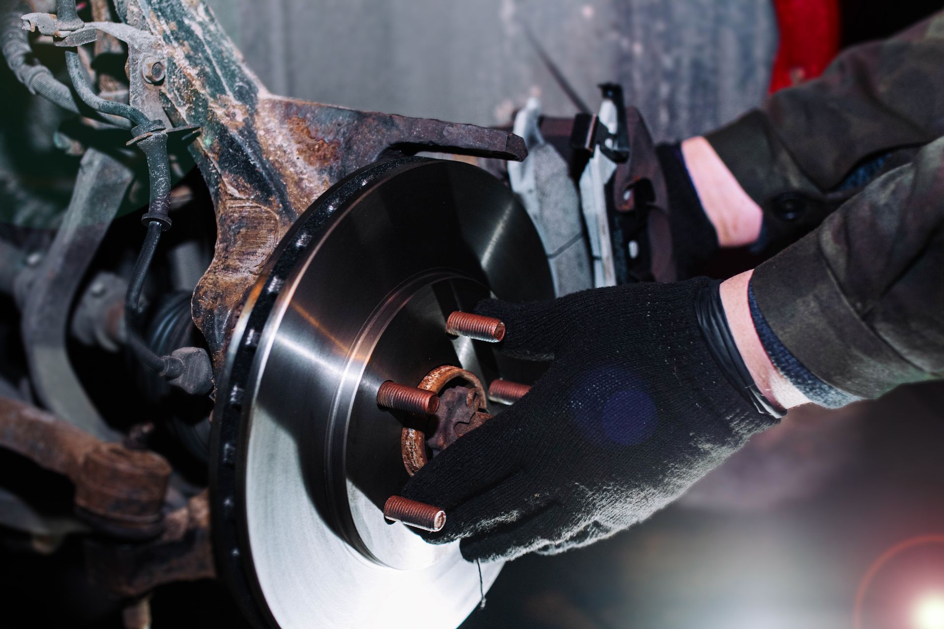 A person wearing black gloves installs a new metal brake disc onto a vehicle's wheel hub in a workshop setting.
