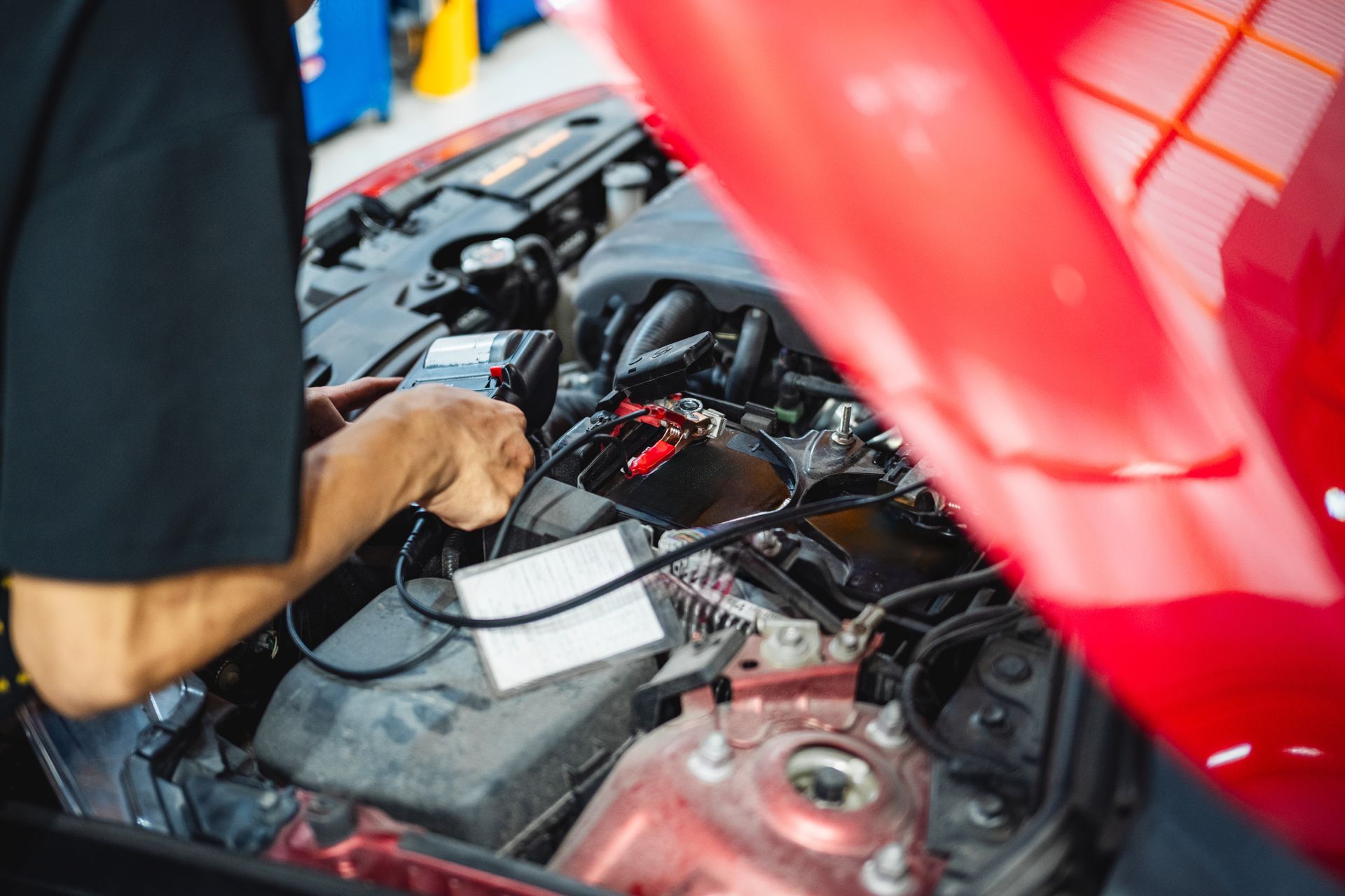 A person wearing a black shirt checks a car battery with a diagnostic tool inside a service garage.